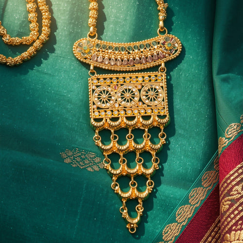 An ornate gold-toned traditional necklace with intricate filigree, circular motifs, and dangling chain elements, displayed on a rich teal-green fabric with a red border.