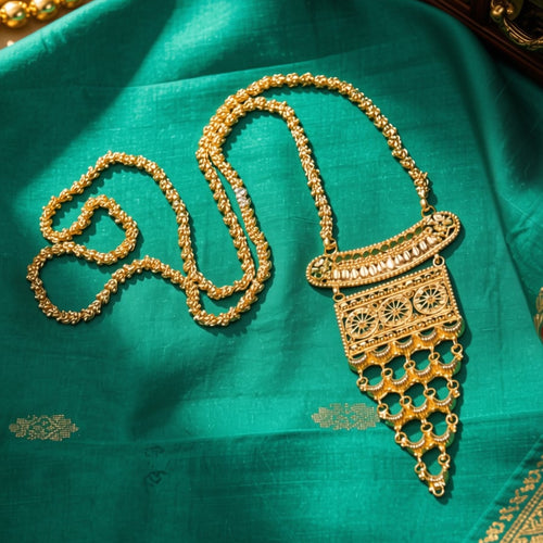 An ornate gold-toned traditional necklace with a detailed pendant, displayed on a rich teal green fabric with decorative props.