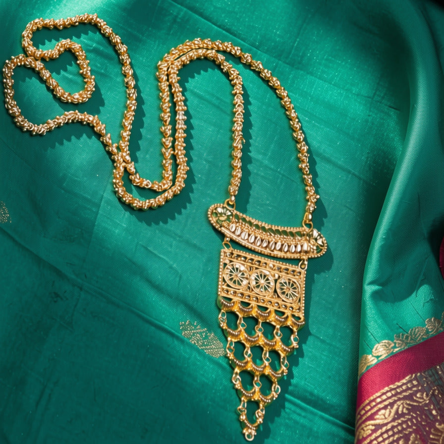 An ornate gold-toned traditional necklace with a detailed pendant, displayed on a rich teal green silk fabric with gold zari borders.