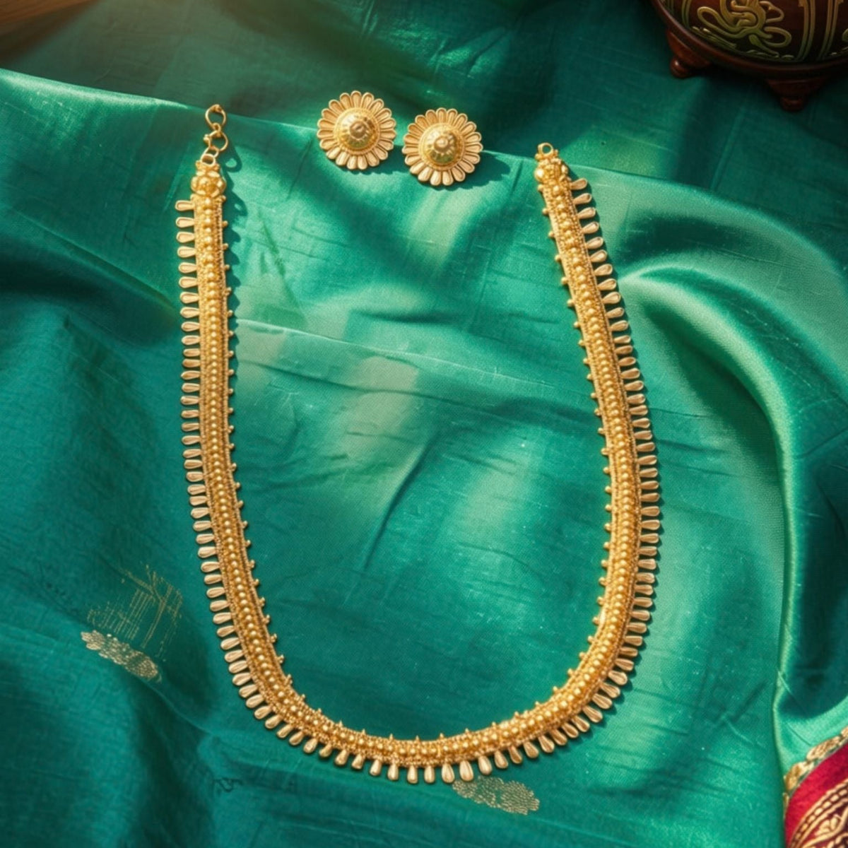 A gold-toned traditional necklace and matching earrings displayed on a rich emerald green silk fabric, with a decorative pot in the background.