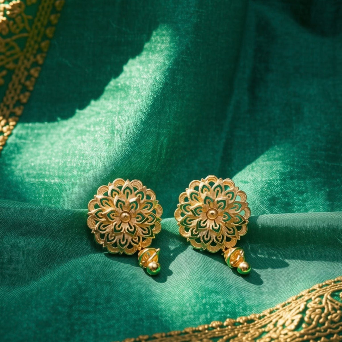 A pair of ornate gold-toned floral earrings with small green enamel accents, resting on a draped emerald green fabric with gold embroidery.