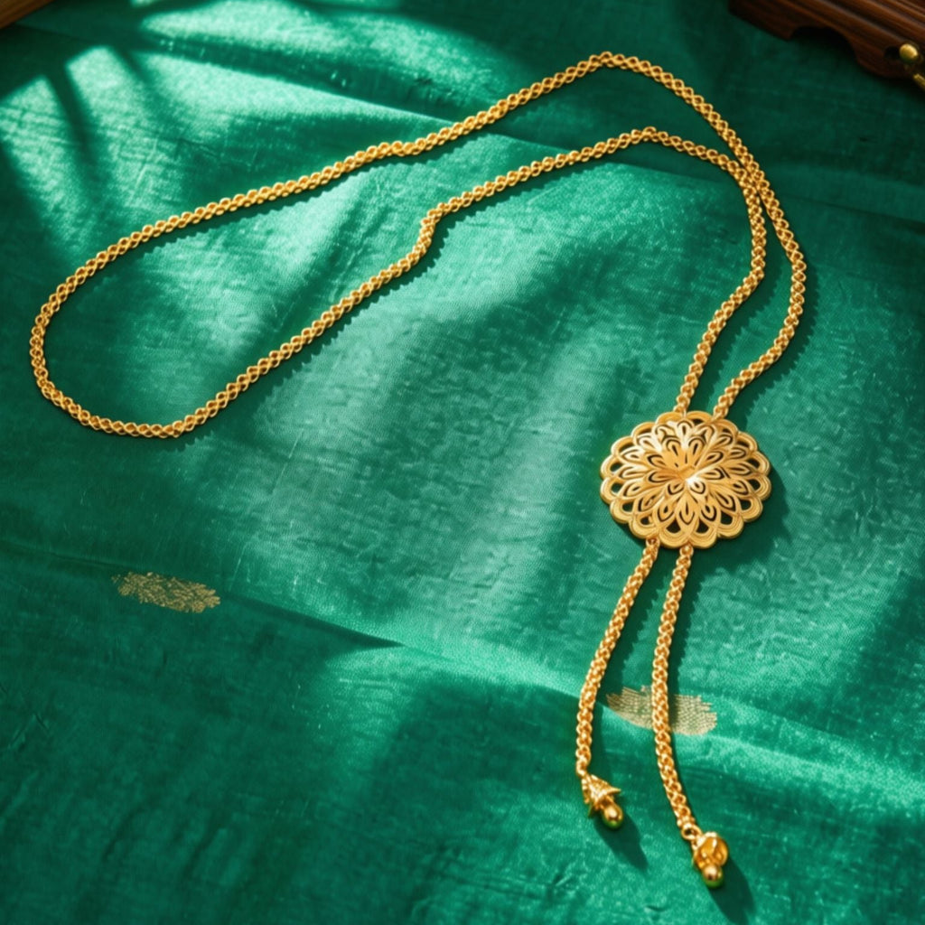 A gold-toned necklace with a circular filigree pendant and long dangling chains, displayed on a textured green fabric surface with warm sunlight and shadows.
