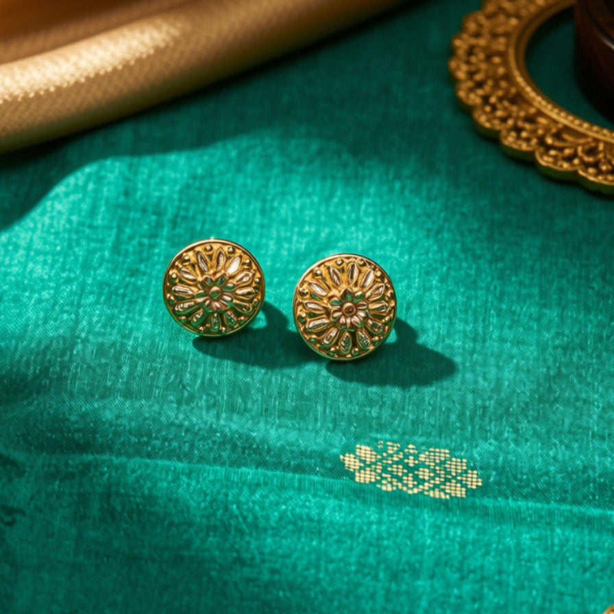 A pair of round gold-toned stud earrings with intricate floral or mandala-like cutout detailing, displayed on a rich teal fabric with gold-toned draped fabric and ornate jewelry props in the background.