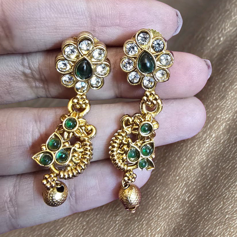 Gold earrings with green gemstones held between fingers on a beige background