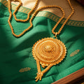 An ornate gold pendant necklace with a large circular, intricately carved pendant and matching chain, displayed on a green embroidered fabric surface.