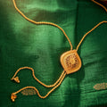 A gold-toned traditional necklace with a square filigree pendant and matching chain, displayed on a rich green fabric with gold motifs.