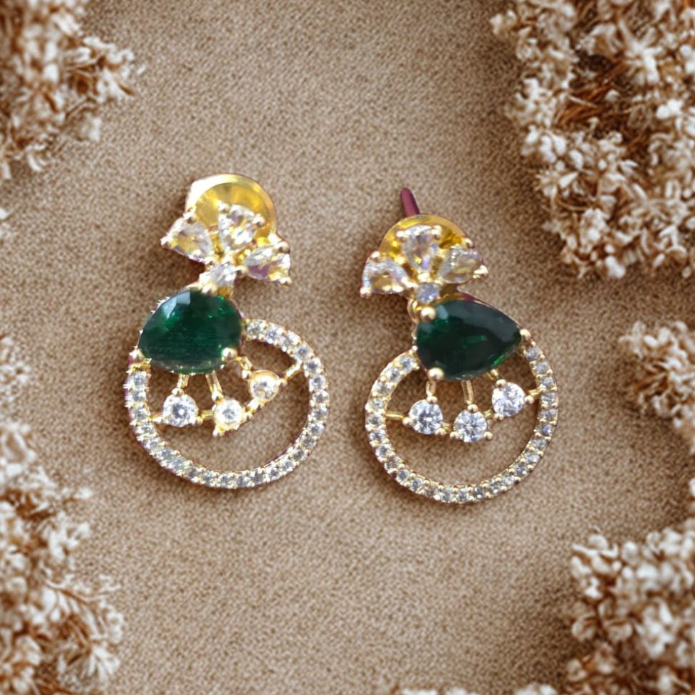 Pair of earrings with green gemstones and gold accents on a beige surface surrounded by dried flowers.